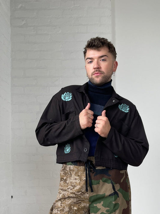 Person wearing a repaired black jacket with green embroidered flowers and camouflage pants against a white wall.